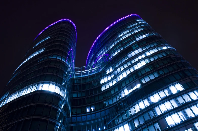 a couple of tall buildings lit up at night, inspired by Zaha Hadid, pexels contest winner, bauhaus, cool purple slate blue lighting, rounded architecture, hannover, purple ribbons