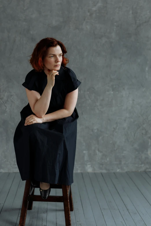 a woman sitting on top of a wooden chair, by Lisa Milroy, wearing a dark dress, woman with red hair, in front of white back drop, bjork