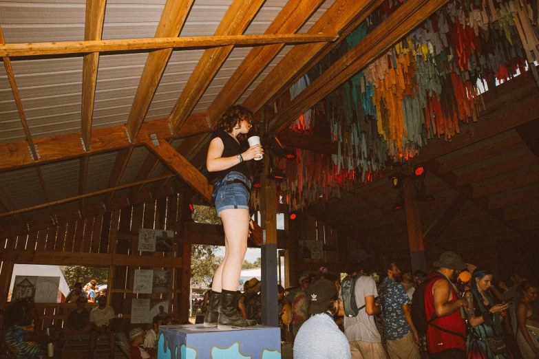 a woman standing on top of a box in a room full of people, unsplash, happening, outside a saloon, tie-dye, covered outdoor stage, ocean dept