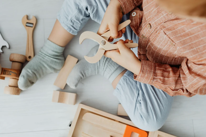 a child sitting on the floor holding a pair of scissors, inspired by Sarah Lucas, trending on pexels, wooden art toys, with mechanical arms that fix it, on a wooden tray, spanners