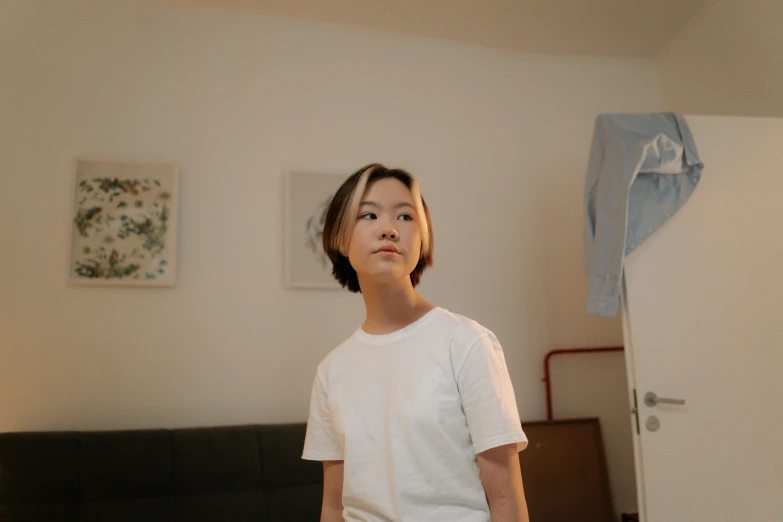 a woman standing in a bedroom next to a bed, by Jang Seung-eop, pexels contest winner, dressed in a white t shirt, boy thin face, young woman looking up, low quality photo