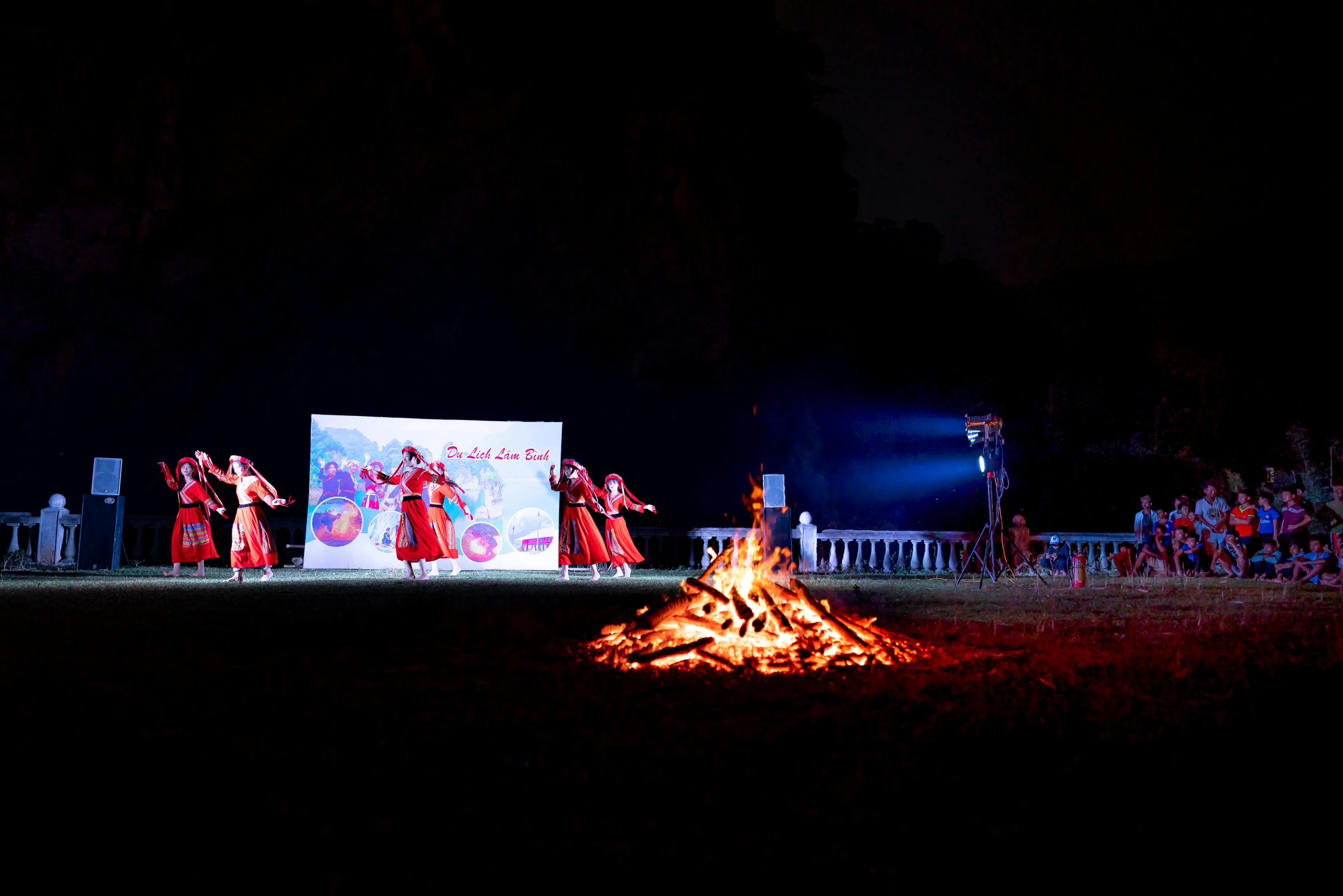 a group of people standing around a fire, interactive art, background: assam tea garden, palace dance, nostalgic 8k, performing on stage