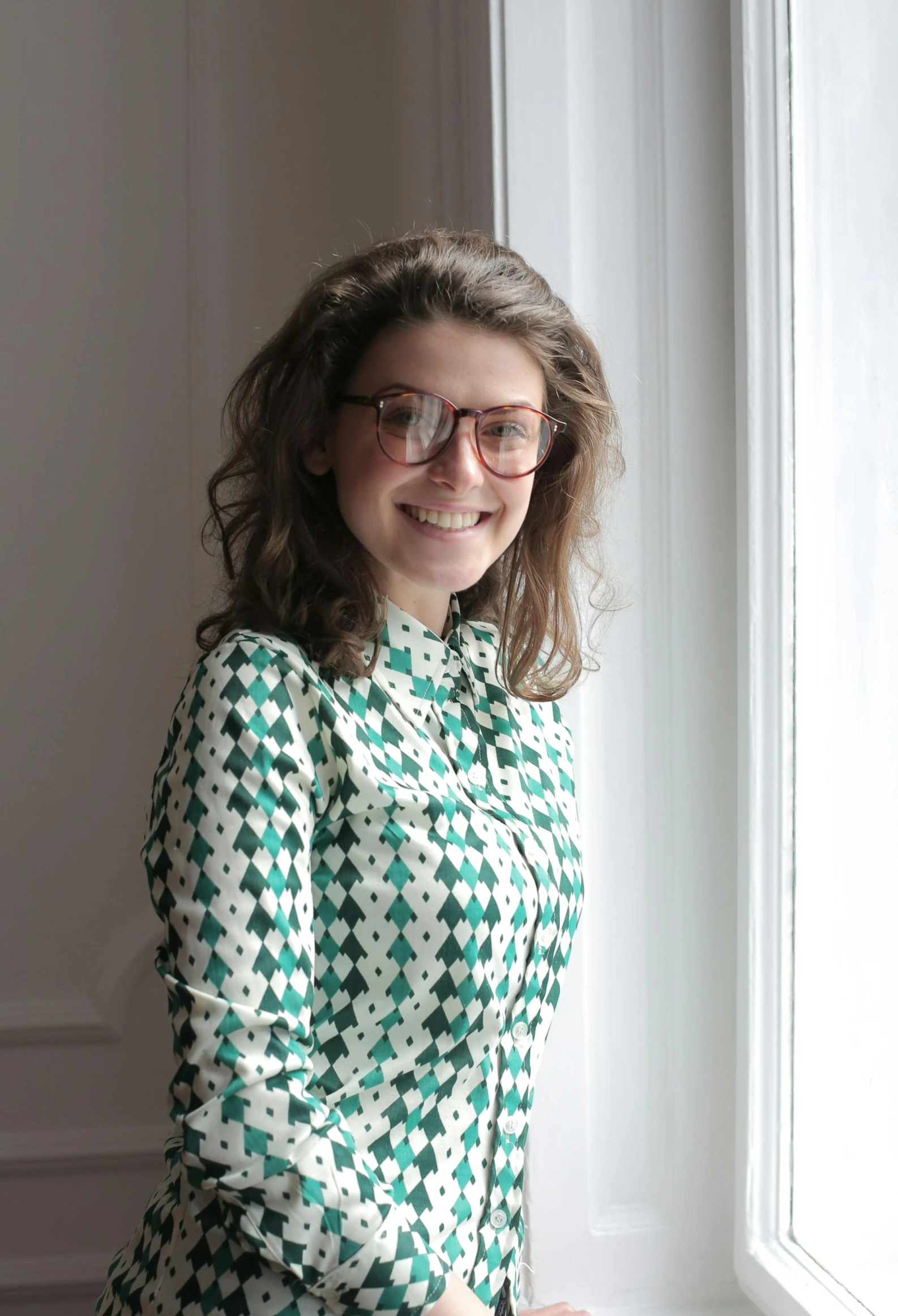 a woman wearing glasses standing in front of a window, a picture, inspired by Anita Malfatti, smiling, patterned clothing, profile image, journalism photo