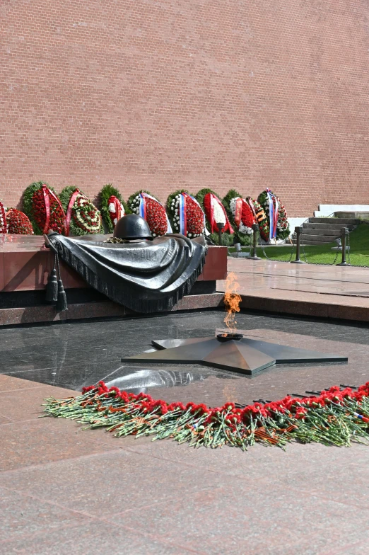 a memorial in front of a building with wreaths around it, red square, fire pit, flower sepals forming helmet, pentagon