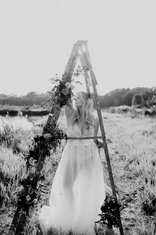 a woman standing in a field next to a ladder, a black and white photo, by Sara Saftleven, unsplash, wearing a wedding dress, flower child, on the altar, promo image