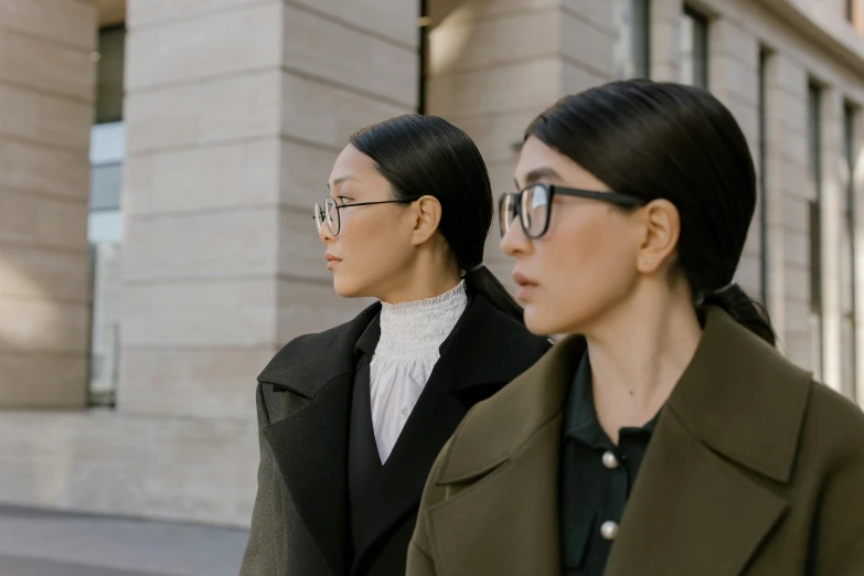 two women standing next to each other in front of a building, inspired by Wang Duo, trending on pexels, square rimmed glasses, wearing black coat, looking from slightly below, ignant