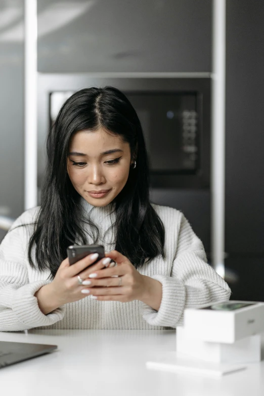 a woman sitting at a table looking at her cell phone, trending on pexels, star trek asian woman, wearing a white sweater, in the kitchen, avatar image