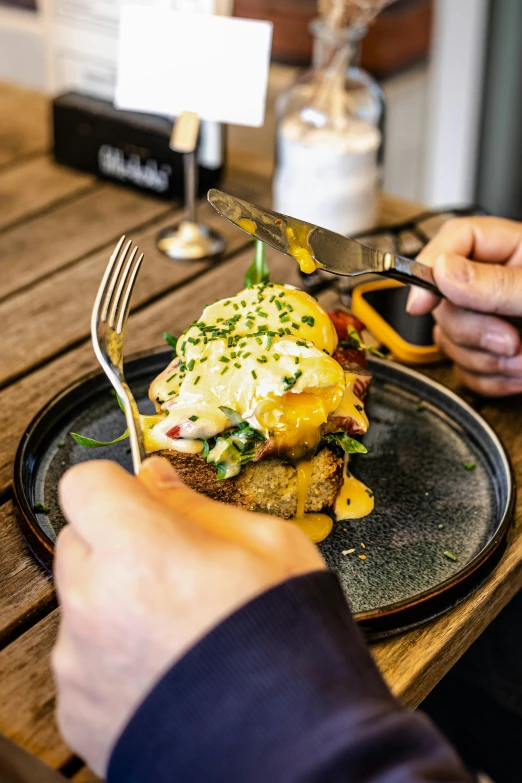 a person sitting at a table with a plate of food, eggs benedict cumberbatch, close up food photography, kete butcher, using fork