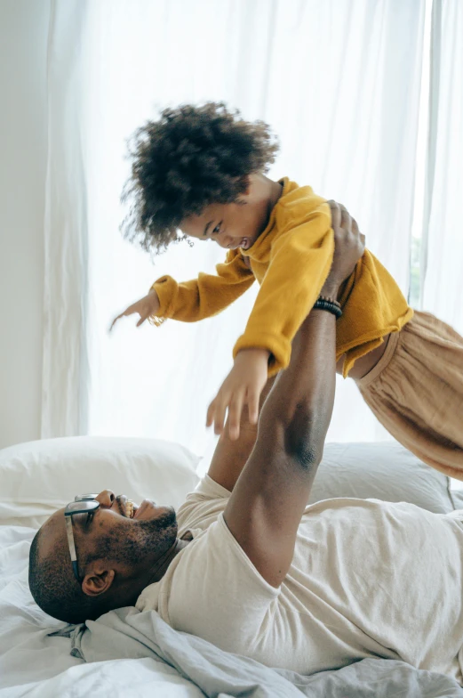 a man laying on top of a bed holding a small child, inspired by The Family Circus, pexels contest winner, african american, arms out, thumbnail, bright daylight