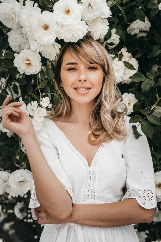 a woman standing in front of a bush of white flowers, inspired by Sydney Carline, pexels contest winner, blonde and attractive features, pokimane, smiling down from above, rose in hair