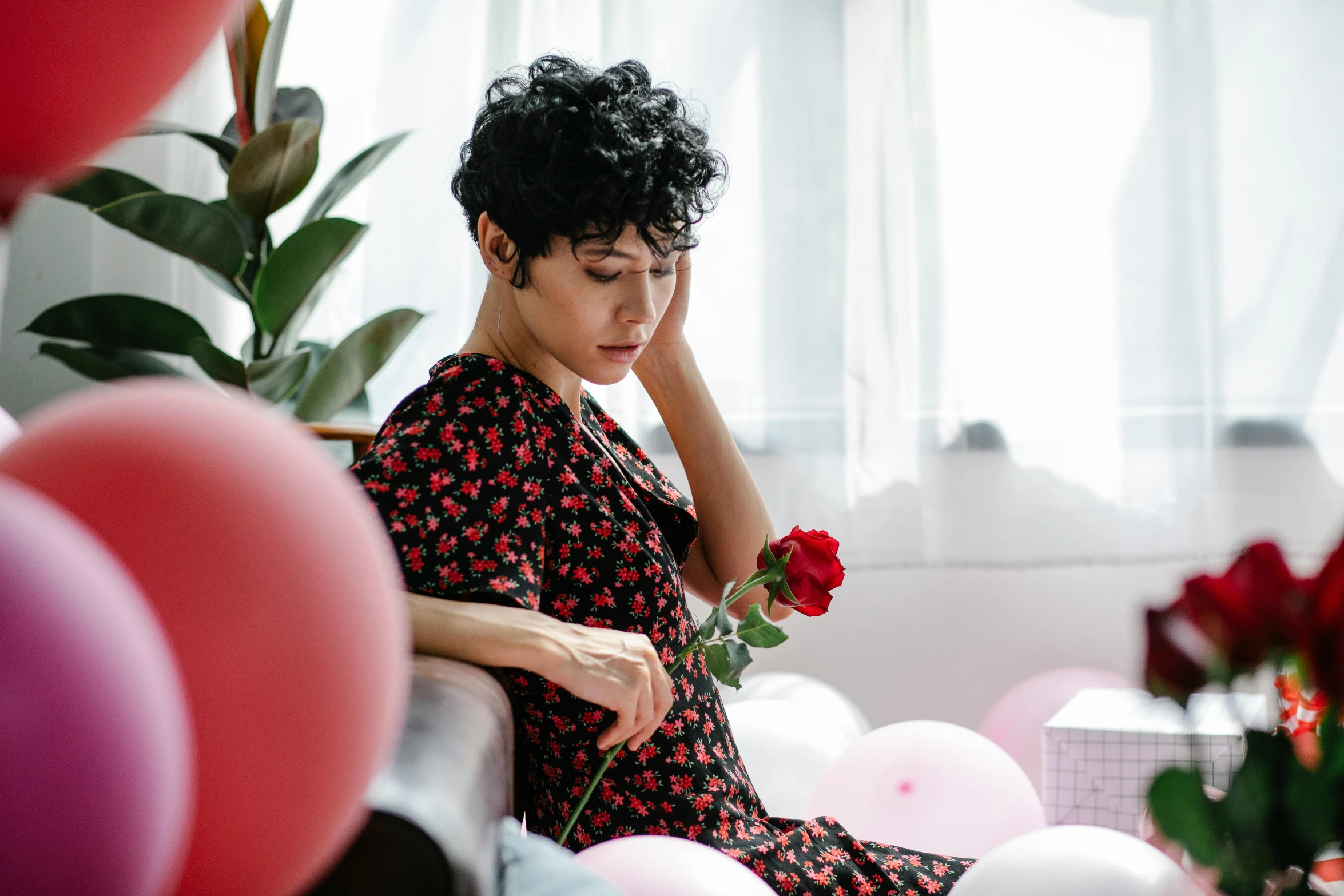 a woman sitting on a couch surrounded by balloons, by Julia Pishtar, pexels contest winner, vanitas, short black curly hair, holding a rose, curly pixie cut hair, sad man