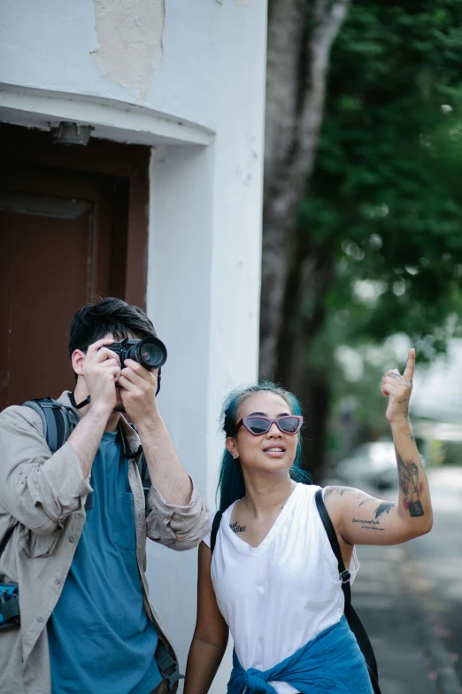 a man taking a picture of a woman with a camera, a photo, by Adam Rex, set on singaporean aesthetic, waving at the camera, wearing shades, tattooed