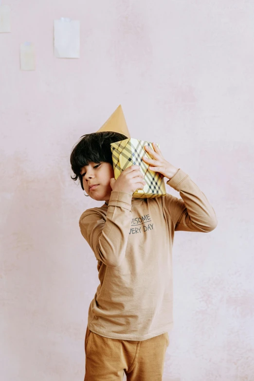 a man standing in front of a pink wall holding a pillow, by Nina Hamnett, pexels contest winner, wearing a party hat, a boy made out of gold, wearing a modern yellow tshirt, brown paper