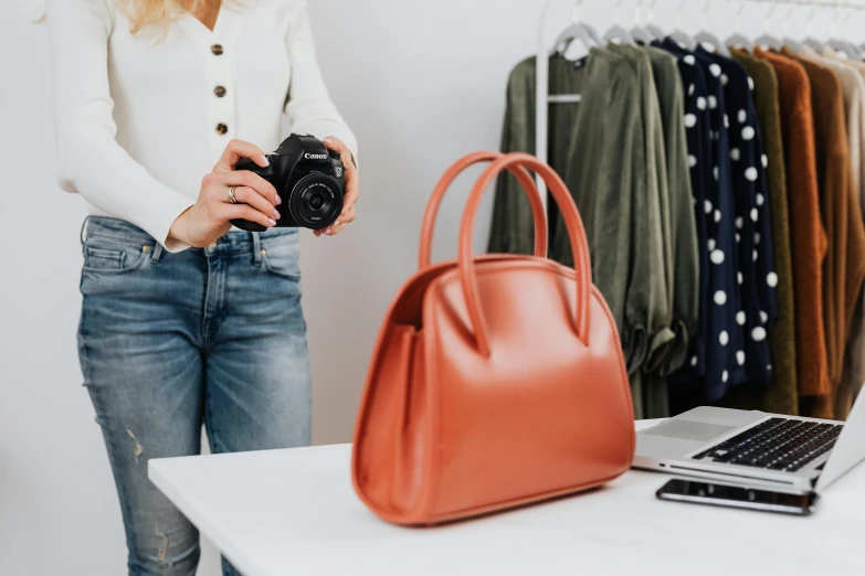 a woman standing in front of a laptop holding a camera, handbag, stores, black and terracotta, at checkout