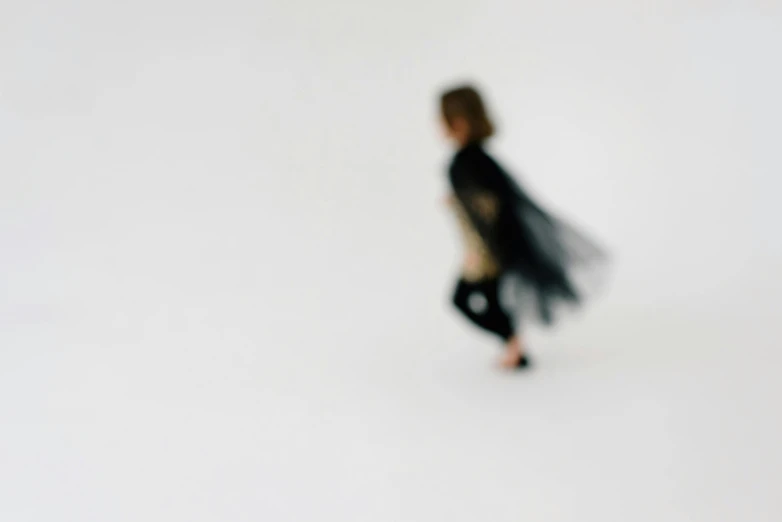 a little girl that is walking in the snow, an album cover, by Emma Andijewska, unsplash, conceptual art, wearing black clothes and cape, in a white room, very blurry, batgirl