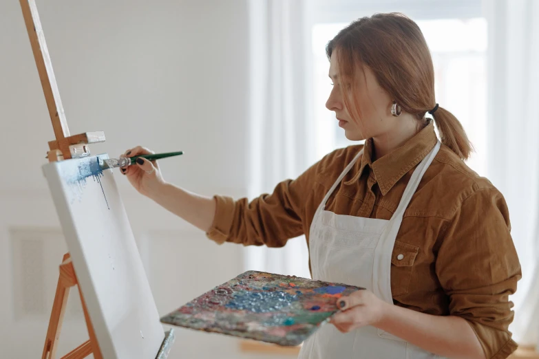 a woman in an apron is painting on an easel, pexels contest winner, white wall coloured workshop, on canvas, looking partly to the left, proportional image