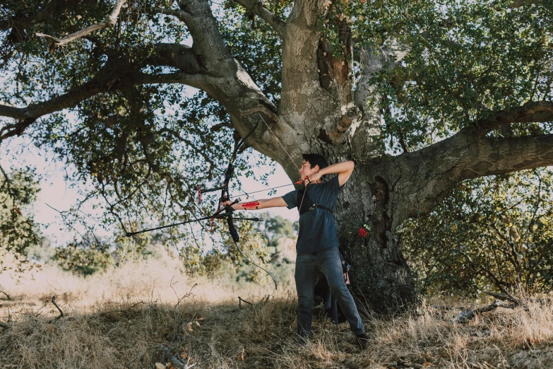 a man holding a bow and arrow in front of a tree, unsplash, renaissance, central california, ignant, behind the scenes photo, background image