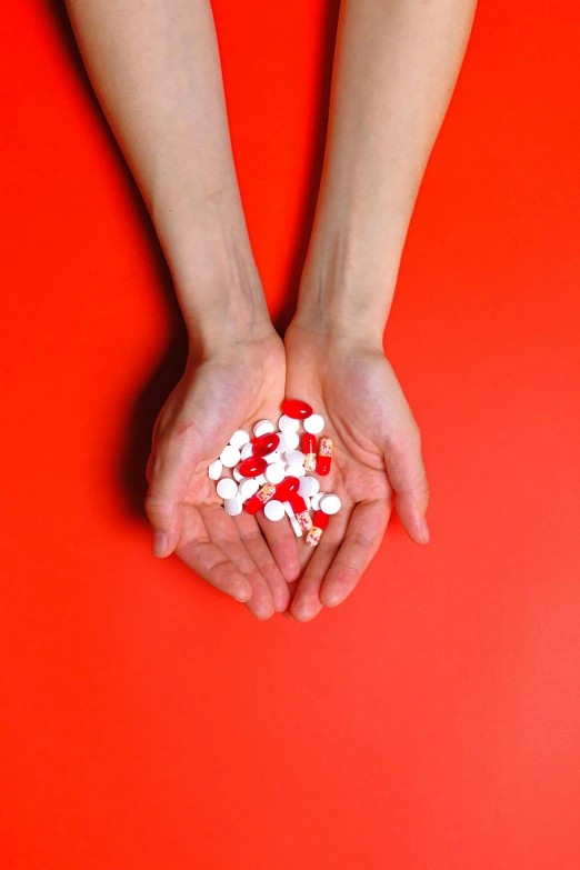 a person holding a bunch of pills in their hands, an album cover, inspired by Damien Hirst, pexels, antipodeans, red and white, vermilion, formulae, shoulder