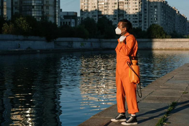 a man in an orange jumpsuit standing next to a body of water, by Julia Pishtar, pexels contest winner, respirator, in an urban setting, pilot outfit, moscow