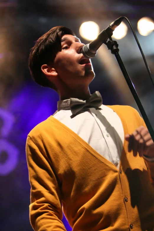 a man in a yellow cardigan singing into a microphone, by Robbie Trevino, bauhaus, bow tie, high quality upload, thumbnail, single subject