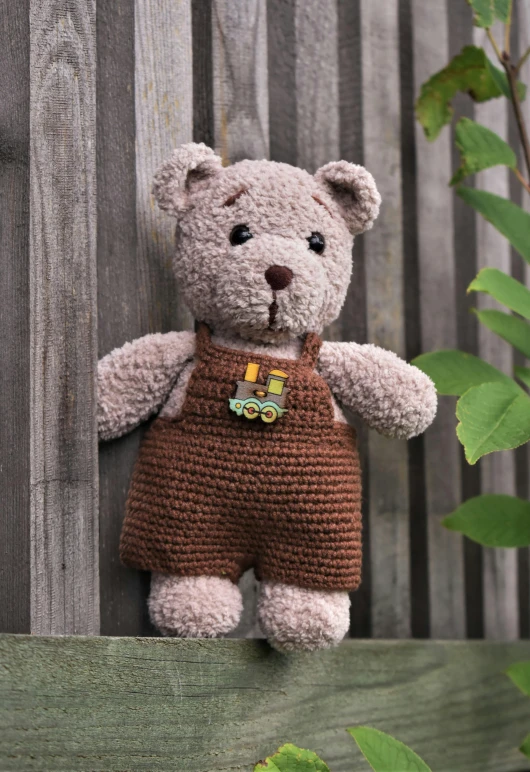 a teddy bear sitting on top of a wooden fence, inspired by Christen Købke, wearing overalls, hughly detailed, crochet, brown