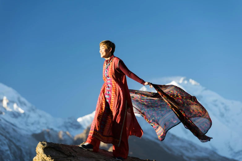 a woman standing on top of a mountain holding a scarf, inspired by Edwin Georgi, happening, wearing a silk kurta, colourful clothing, embroidered robes, avatar image