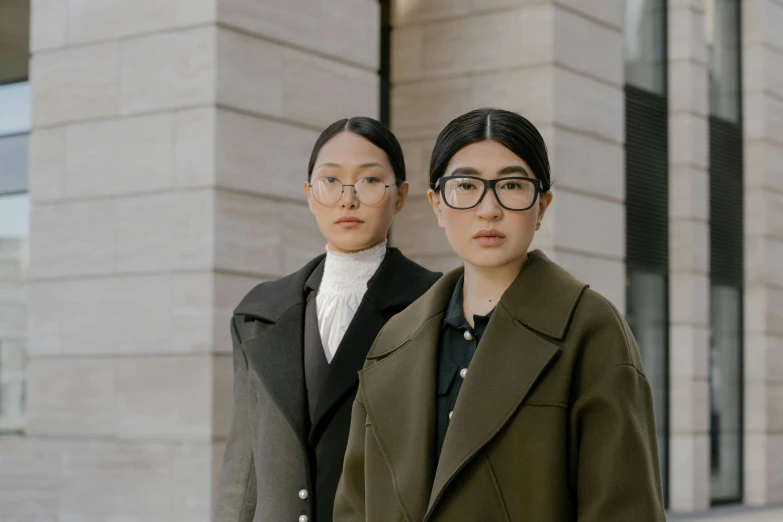 two women standing next to each other in front of a building, inspired by Wang Duo, trending on pexels, square glasses, coat, looking at viewer, korean symmetrical face