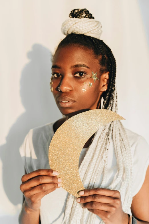 a close up of a person holding a hat, inspired by Ras Akyem, afrofuturism, glitter makeup, shining crescent moon, cardboard cutout, girl with plaits