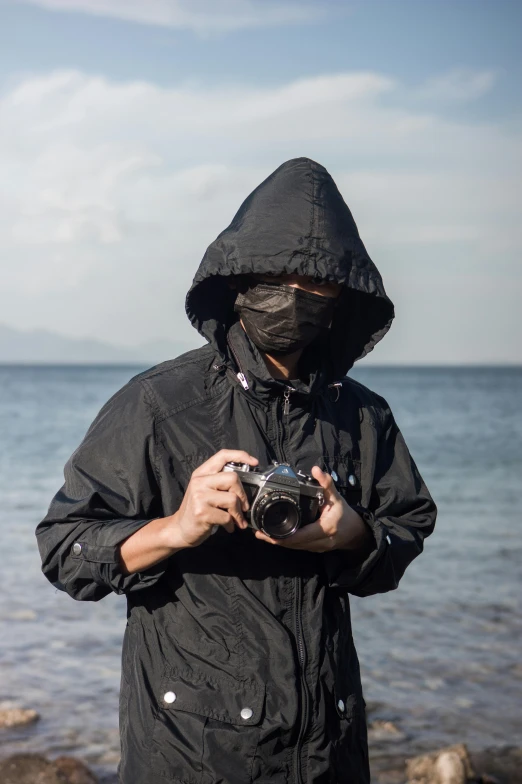 a man standing on a beach holding a camera, inspired by Kanō Hōgai, wearing a dark hood, wearing bandit mask, reflective suit, candid photography