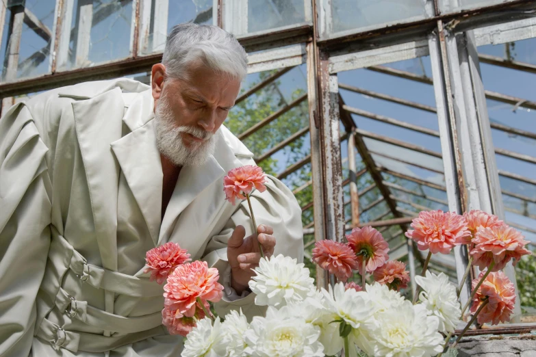 a man looking at flowers in a greenhouse, inspired by Cecil Beaton, overalls and a white beard, wearing robes of silver, promotional image, video still