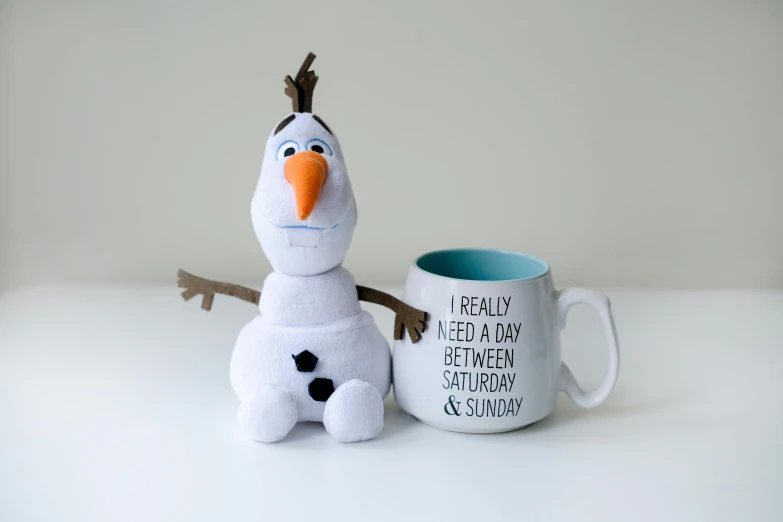 a stuffed snowman sitting next to a coffee mug, a cartoon, inspired by Olaf Rude, happening, on a white table, elsa from frozen, i és complex, good day