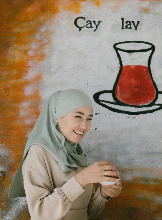 a woman standing in front of a wall with a painting on it, a picture, inspired by Maryam Hashemi, pexels contest winner, hurufiyya, is ((drinking a cup of tea)), malaysian, subject is smiling, islamic