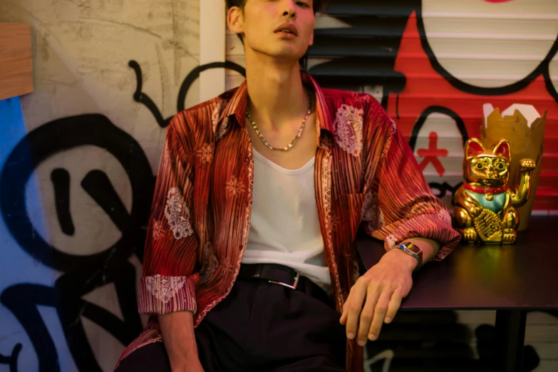 a man sitting at a table in front of a wall with graffiti, an album cover, trending on pexels, renaissance, classy yukata clothing, red jewelry, wonbin lee, wearing stripe shirt