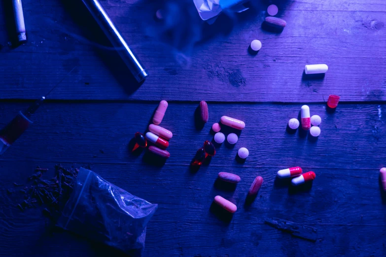a bunch of pills sitting on top of a wooden table, a still life, by Julia Pishtar, unsplash, process art, blue and purple lighting, meth lab, light red and deep blue mood, black and blue and purple scheme