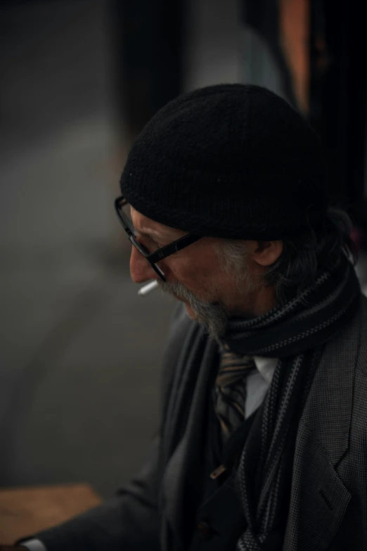 a man sitting at a table with a cigarette in his mouth, a character portrait, pexels contest winner, under a gray foggy sky, yohji yamamoto, on the sidewalk, man wearing a closed cowl
