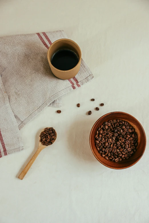 a cup of coffee next to a bowl of coffee beans, a still life, inspired by Ceferí Olivé, trending on unsplash, coffee stain on napkin, spoon placed, detailed product image, detail shot