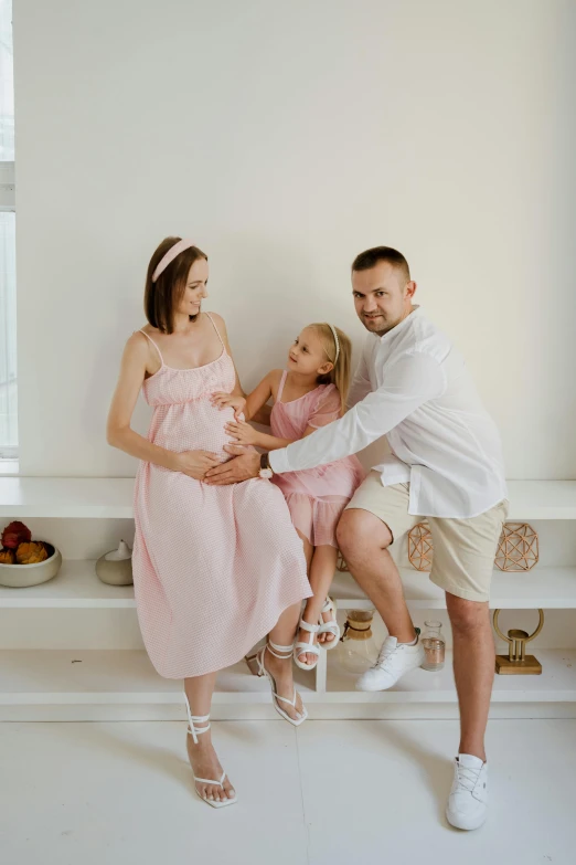 a couple of people that are sitting on a bench, white and pink cloth, with a kid, in white room, pregnant belly