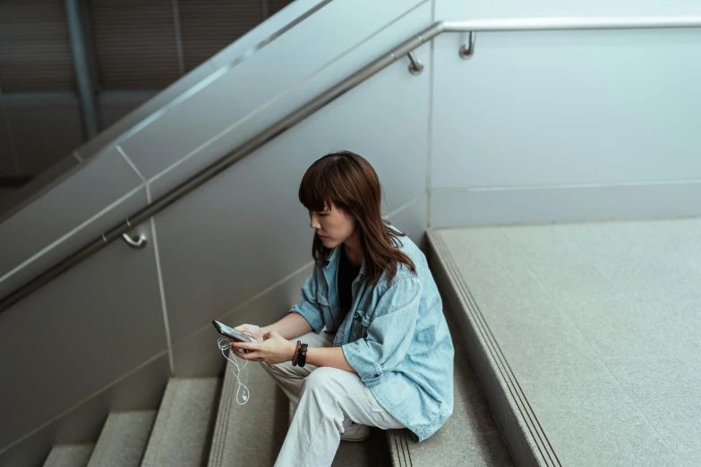 a woman sitting on a set of stairs using a cell phone, trending on pexels, wearing a light blue shirt, sakimichan, gaming, androgynous person