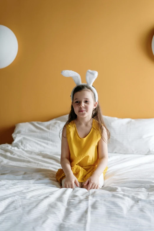 a little girl in bunny ears sitting on a bed, inspired by Elsa Beskow, pexels contest winner, pop art, yellow orange, hotel room, easter, minimalissimo