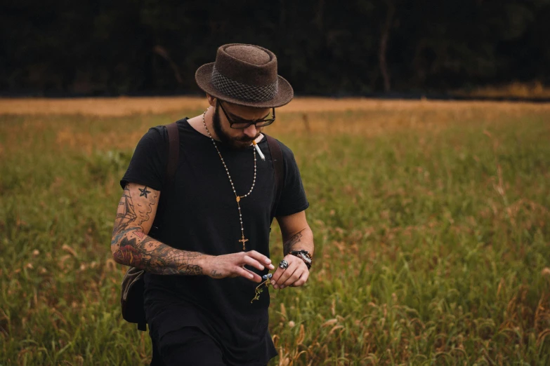 a man standing in a field looking at his cell phone, a portrait, by Adam Marczyński, pexels contest winner, tattooed, tipping his fedora, beads cross onbare chest, wearing a black t-shirt