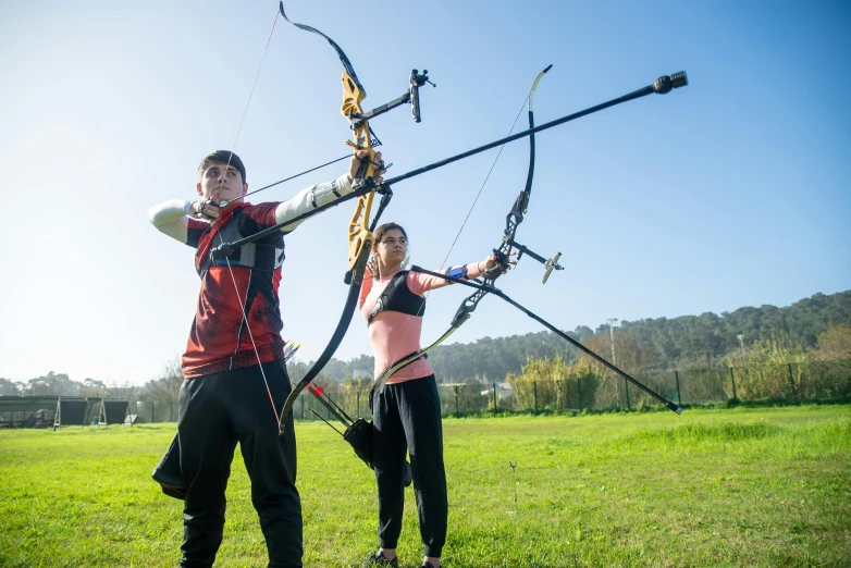 a couple of people standing on top of a lush green field, aiming a bow and arrow, local gym, manuka, avatar image