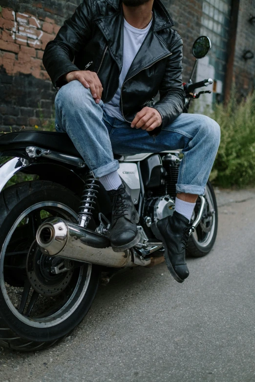 a man sitting on a motorcycle in front of a brick wall, by David Donaldson, trending on unsplash, heavy boots, sitting on a store shelf, leather cuffs around wrists, wearing denim