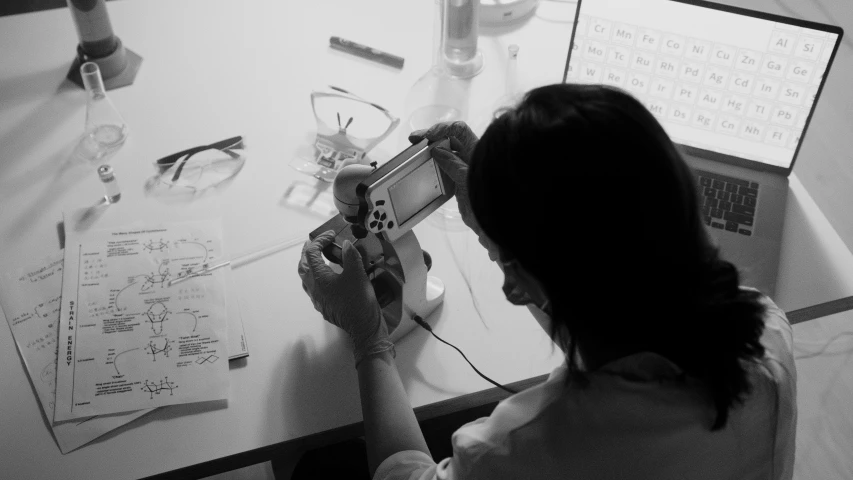 a woman sitting at a desk in front of a computer, a black and white photo, process art, nintendo ds, plating, maintenance, holding controller