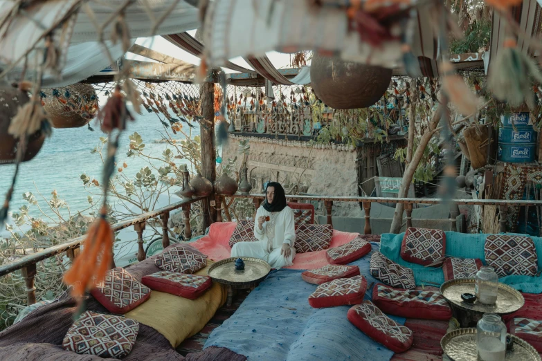 a woman sitting on top of a bed next to the ocean, maximalism, inside an arabian market bazaar, cottagecore hippie, coffee shop, photo”