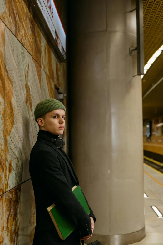 a man leaning against a wall in a subway station, an album cover, inspired by Anna Füssli, unsplash, bauhaus, green hat, nonbinary model, ukrainian, beanie