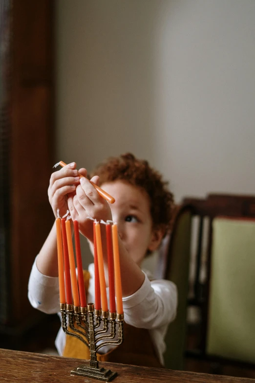 a little girl sitting at a table with a hanukah menorah, by Jessie Algie, pexels contest winner, crayon art, orange fluffy spines, drawing an arrow from his quiver, instagram photo, orange light