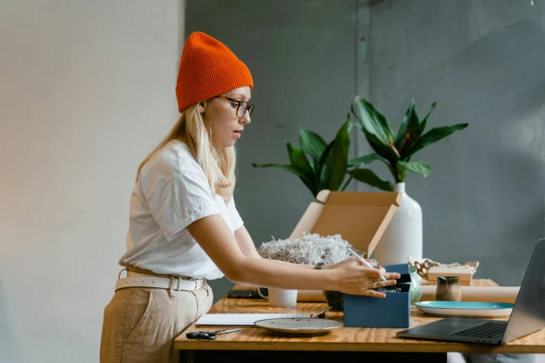 a woman sitting at a table working on a laptop, by Julia Pishtar, trending on pexels, arbeitsrat für kunst, wearing a beanie, delivering parsel box, gray and orange colours, female gnome artificer