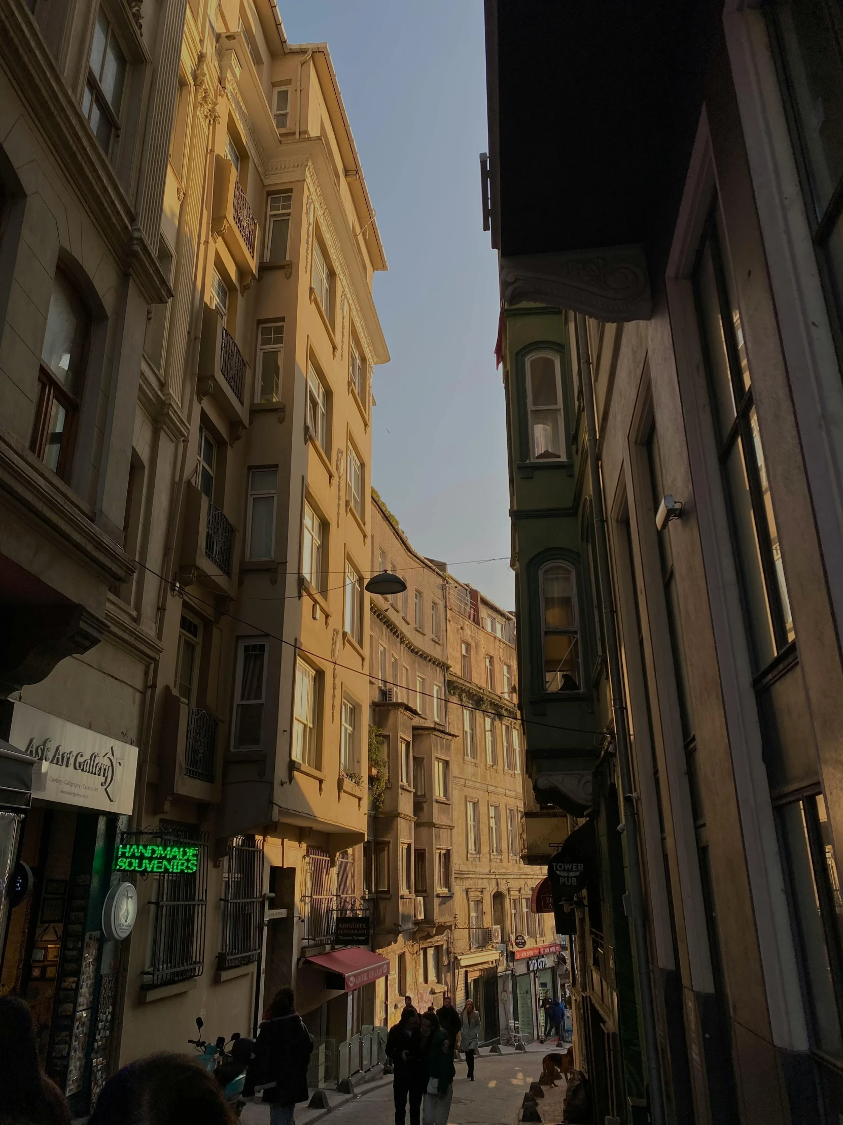 a group of people walking down a street next to tall buildings, a picture, inspired by Niyazi Selimoglu, pexels contest winner, art nouveau, humid evening, view from inside, preserved historical, soft sunlight dappling