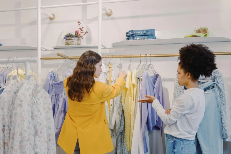 a couple of women standing in front of a rack of clothes, trending on pexels, renaissance, calmly conversing 8k, lady using yellow dress, diverse, advanced technique