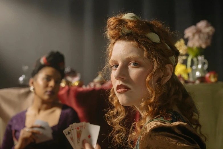 a woman holding a playing card next to another woman, inspired by Hans Holbein the Elder, renaissance, 2 0 2 1 cinematic 4 k framegrab, redhead queen in heavy red armor, person in foreground, colour photograph